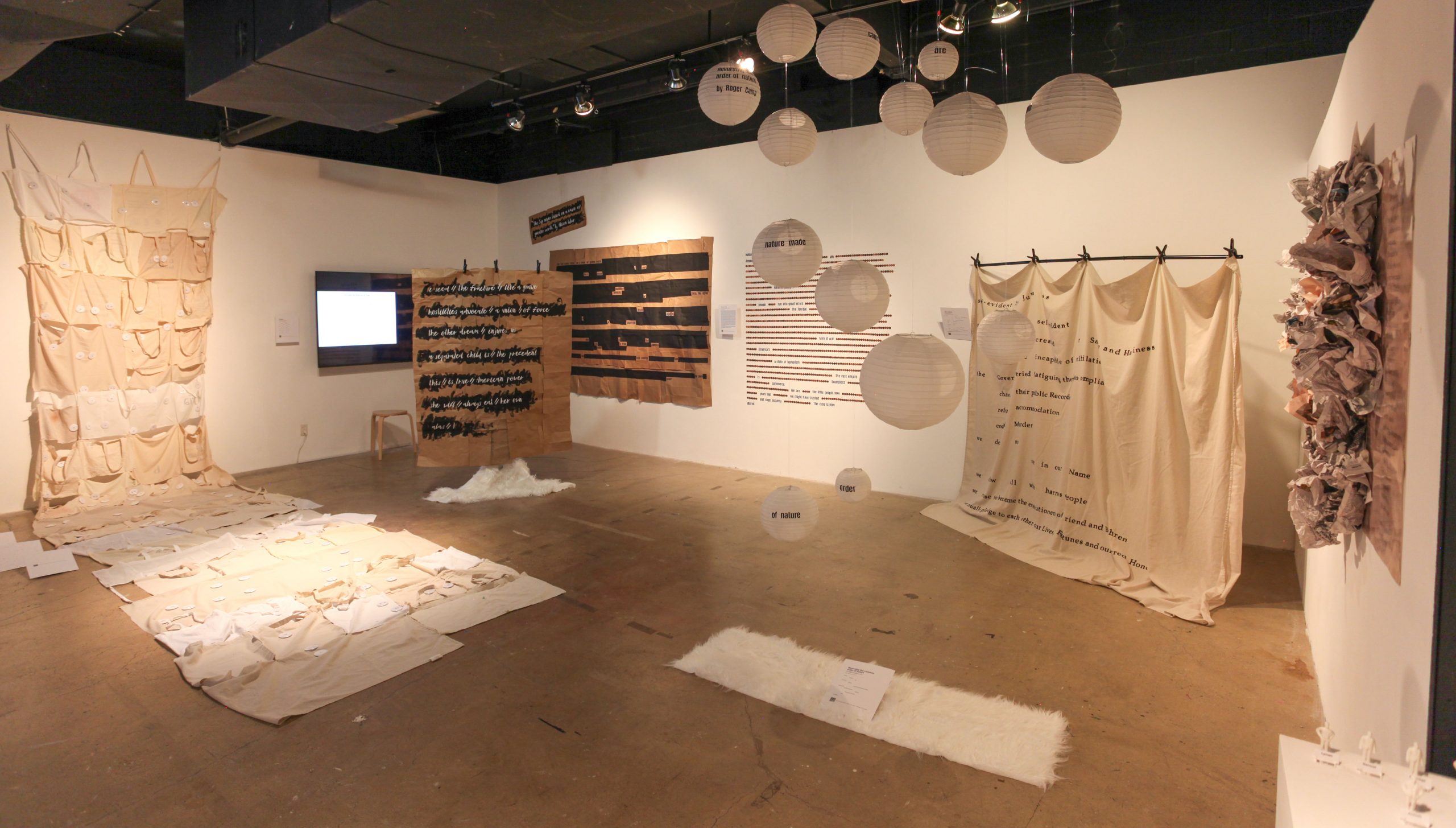 Wide view of the poetry gallery installation featuring large-scale textile and mixed-media works in neutral beige, cream, and brown tones. Fabric panels with stitched pockets, printed text, and layered materials hang on the walls, while suspended white paper lanterns with words printed on them float overhead. A glowing video screen and wooden stool sit in the back corner. Pieces of fabric and paper are spread across the floor, alongside small white rugs and sculptural elements, creating an immersive, contemplative exhibition environment.