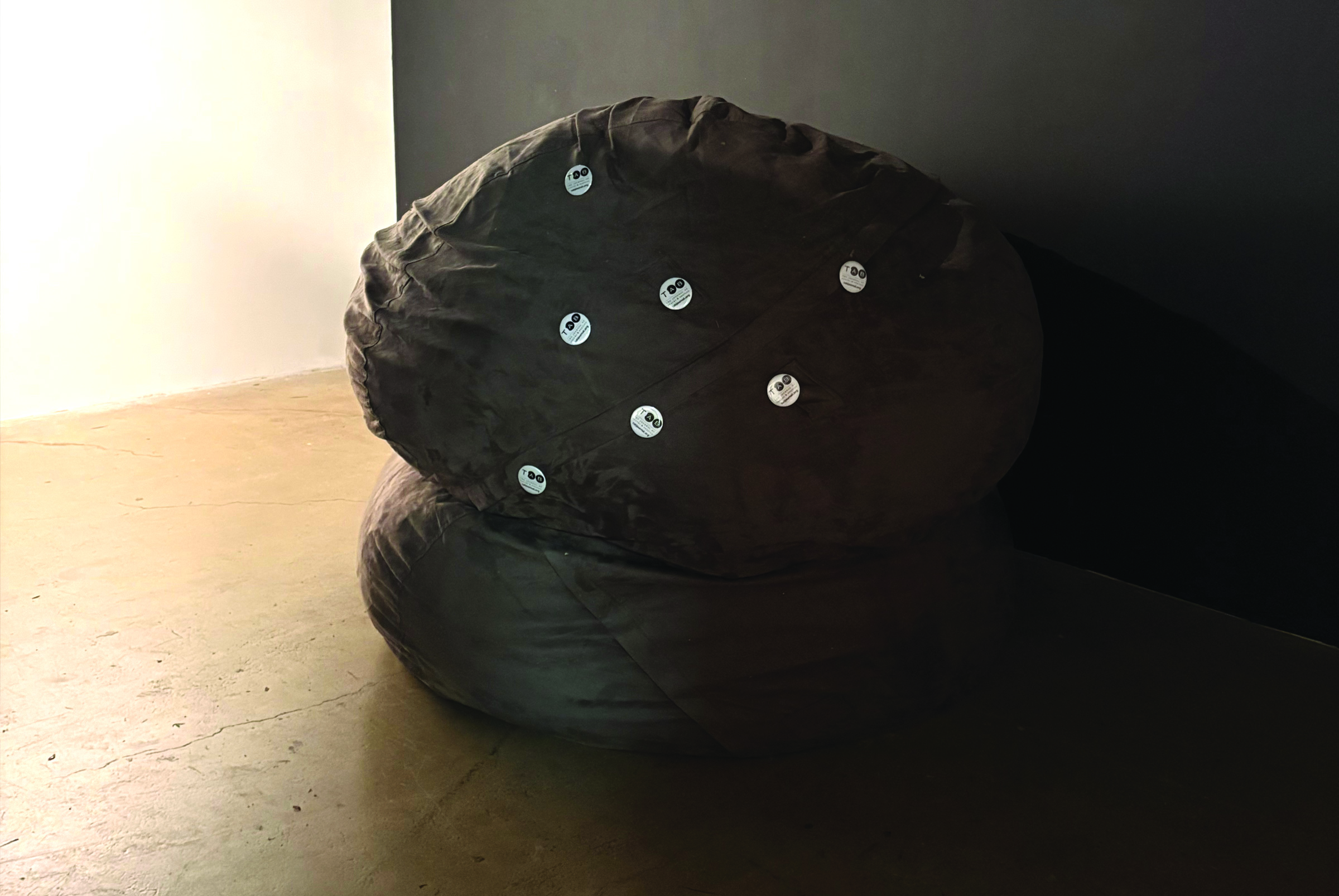 A dark brown, deflated or slouchy fabric pouch or cushion lying on a wooden surface, with several small silver metal grommets or snaps attached to its surface
