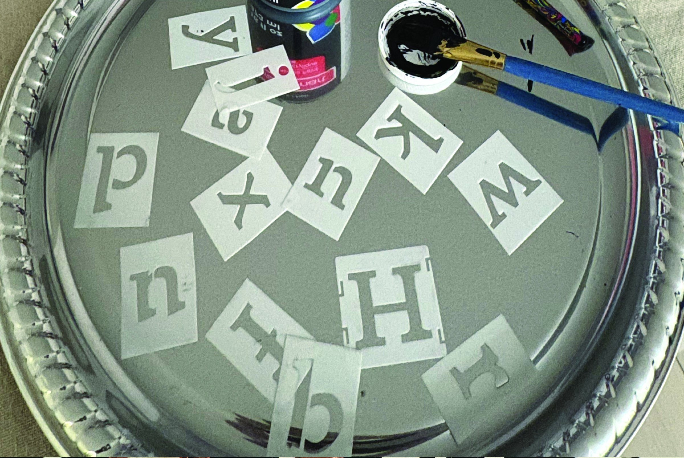 A round silver tray holding loose white letter stencils of various sizes and fonts, scattered across the surface. A small can of black paint, a paintbrush, and other art supplies are visible at the top of the tray.