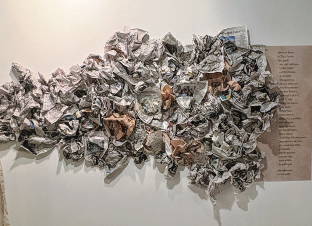 A wall-mounted sculpture made of densely crumpled and layered newspaper or printed paper, forming a large organic, cloud-like mass. Printed text is visible on the wall beside it.