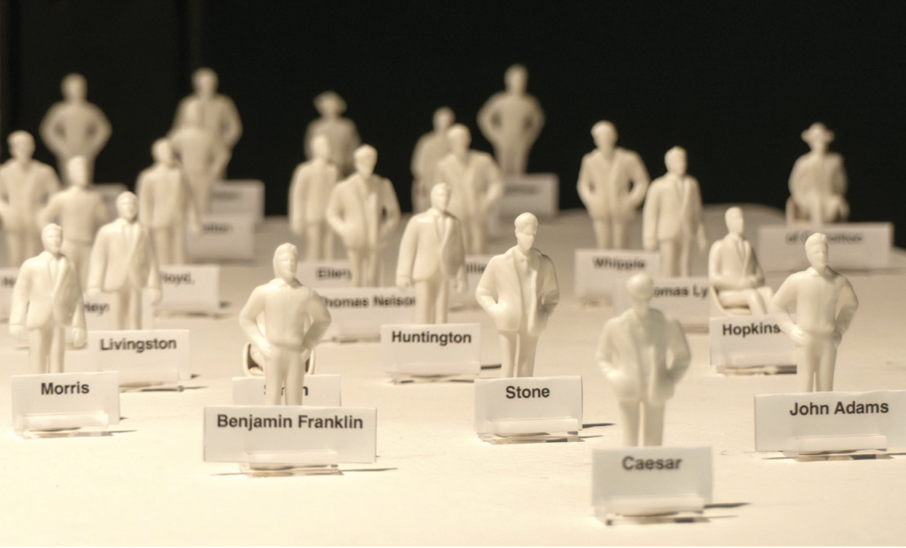 Numerous small white figurines of human forms standing in rows on a surface, each labeled with a name tag. Visible names include Benjamin Franklin, Morris, Livingston, Huntington, Hopkins, Stone, Caesar, and others.