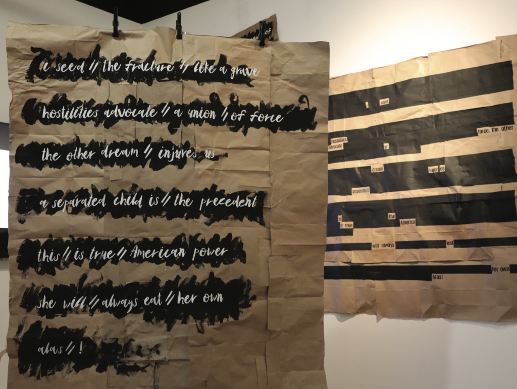 Large sheets of brown kraft paper hung on a wall, painted with bold black brushstroke-style bands. Over the brushstrokes, handwritten white script poetry is visible, with lines including phrases such as 'hostilities advocate a union of force' and 'a separated child is the precedent.